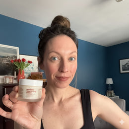 Woman holding a jar of skincare cream in a room with blue walls and decor.